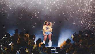 Beyoncé standing on stage in a yellow sweatshirt, denim cutoff shorts, and white fringe boots. She is holding a microphone in her right hand and has her left resting on her hip. There is a marching band sitting on risers below her all wearing matching yellow uniforms and holding their instruments. Behind Beyoncé there are sparks falling from the rafters above her.