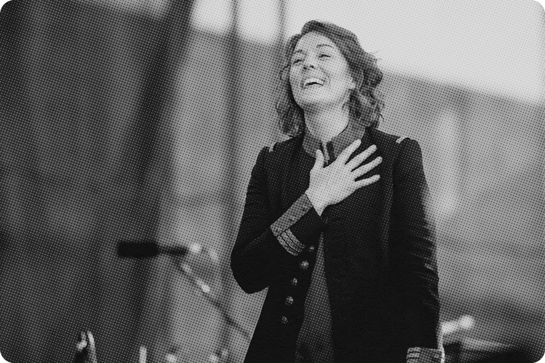 A black and white photo of Brandi Carlile standing on stage smiling outward with her right hand placed over her heart. She is wearing a large jacket with buttons down the front, a thick collar, and large cuffs and buttons around the wrists. She is on stage and behind her is a microphone.
