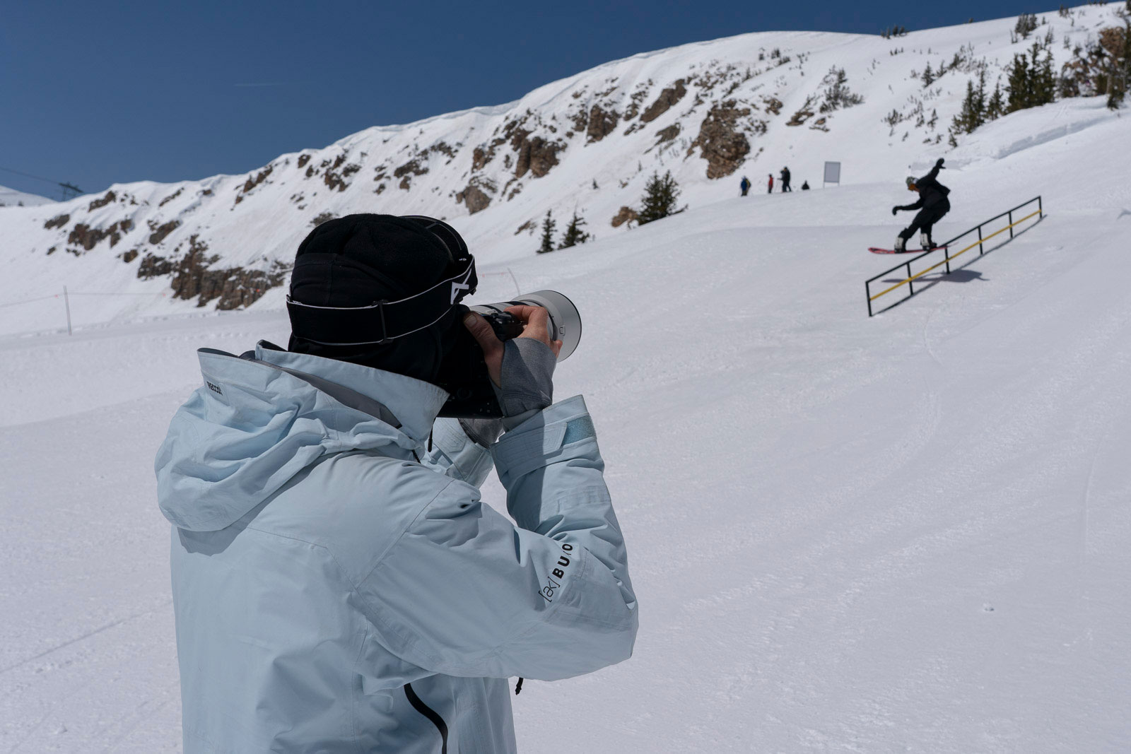 person taking picture on ski hill of person snowboarding on rail in terrain park