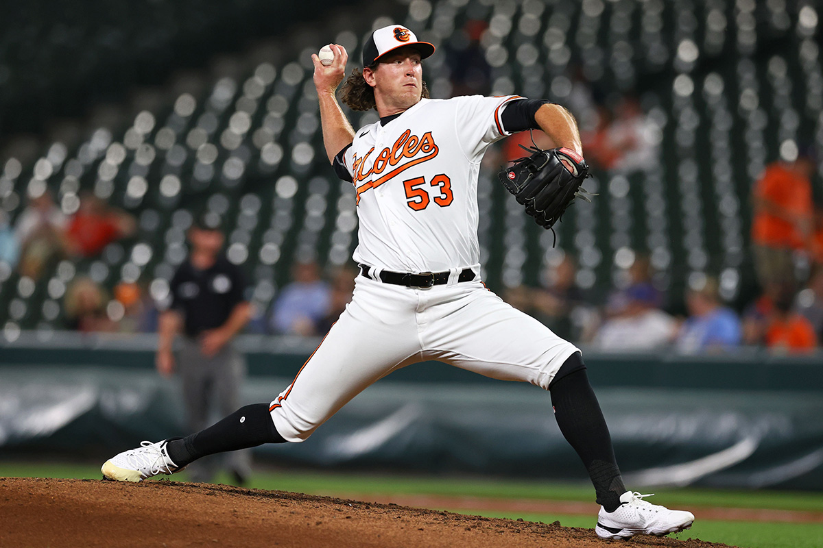 Action shot of Baltimore Orioles pitcher Mike Baumann stretched out mid-pitch with the ball in his right hand. Mike is wearing the classic white, orange and black Baltimore Orioles uniform.