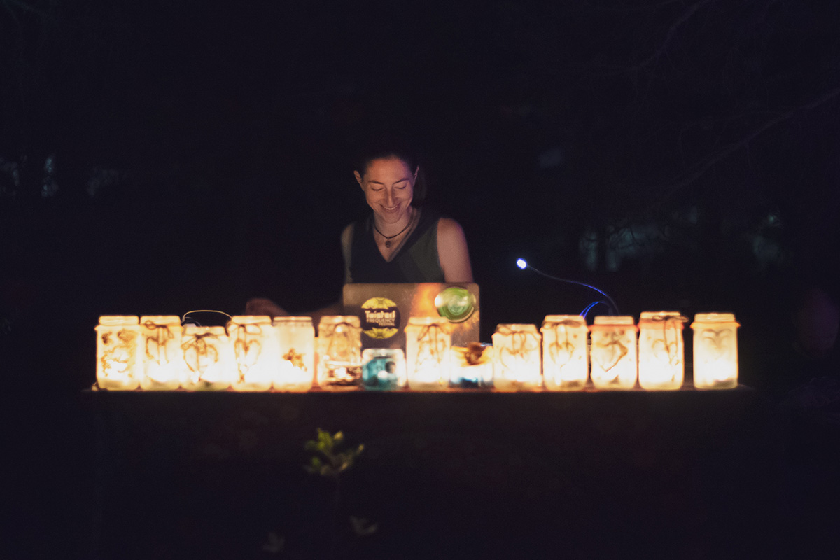Photo taken in the dark of Sara Niksic making music on her capturer at a desk. A row of mason jar glasses are lined up across the desk and are filled with yellow tea lights that are shining on Sarah's face.