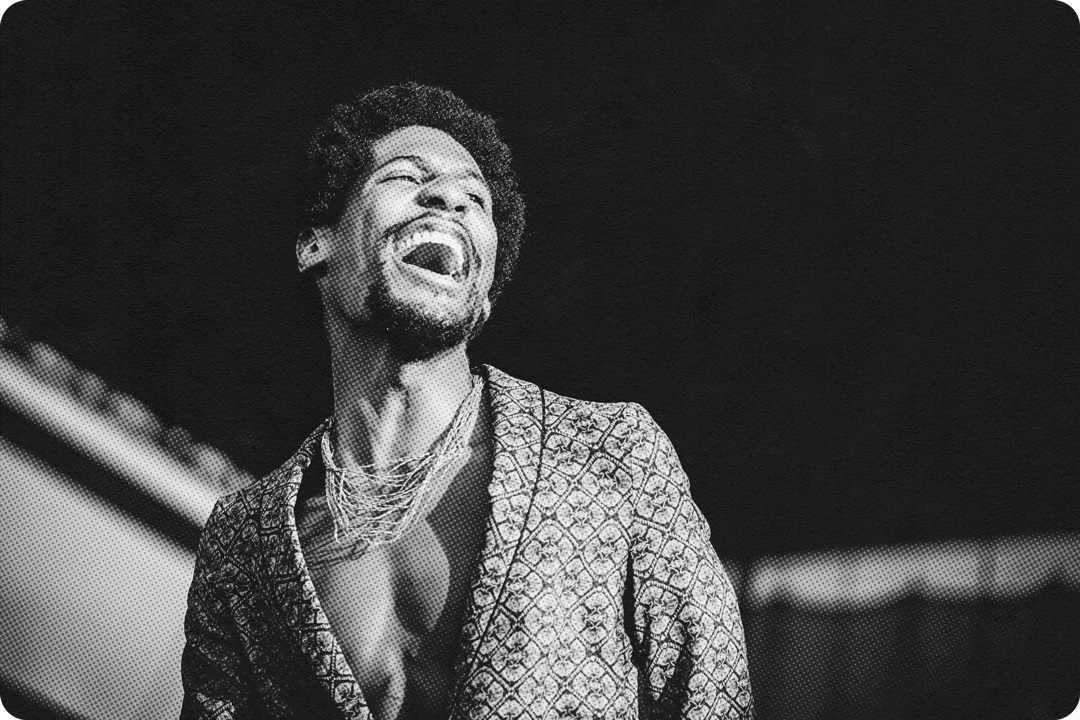 Jon Batiste smiling while on stage. The photo is taken from the chest up, Batiste is wearing a patterned blazer with no shirt underneath and a tangle of necklaces around his neck. He has an open mouthed smile and is looking up to the right of the frame.
