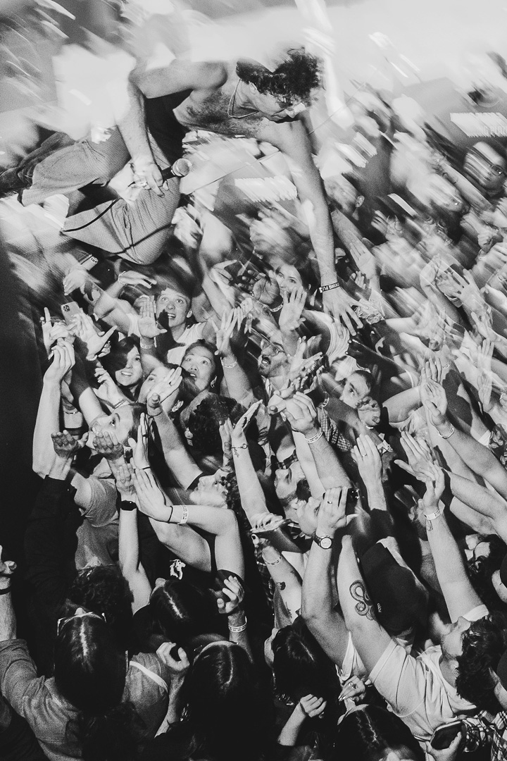 Black and white photo of a man in the middle of a jump to crowd surf. The man jumping is not wearing shirt but is holding a wired microphone. The crowd below him all has their hands up in preparation to catch him. Around the top of the photo is lots of motion blur as the camera attempts to capture the high energy moment.