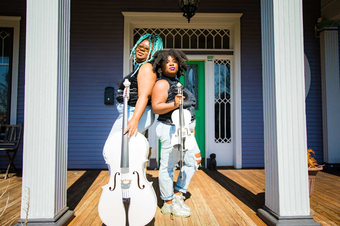 Two women posing for a photo together on the front porch of a victorian style house. The women are standing back to back with their faces turned to their side to face the camera. They are both wearing jeans and black tank tops but the woman on the left is holding up a white cello and the woman on he right is holding up a white violin and bow.