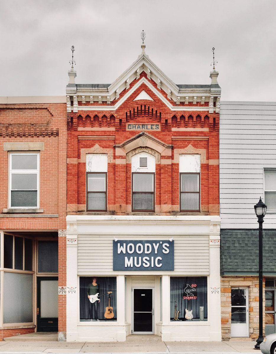 Storefronts with a red brick intricate patterned brick work with a blue sign "Woody's Music" in the center. In the windows are a mannequin with guitar and a "go sharks" neon sign. 