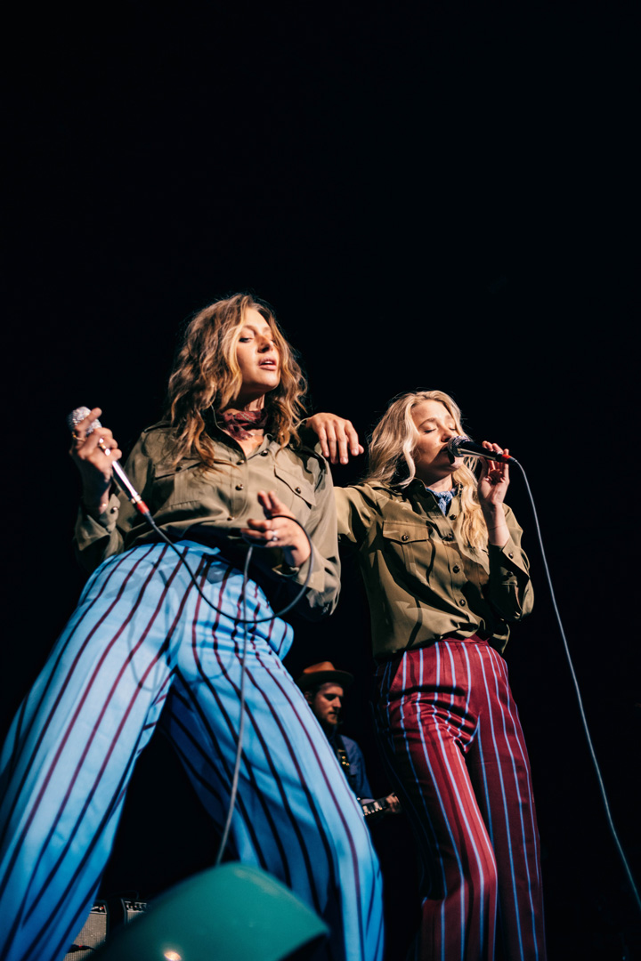 Two women singing on stage together, the one on the left has the microphone in her right hand and the cord in the other while the girl on the right is holding the microphone to her mouth with one and and using the other to lean on the other girl's shoulder. The girl on the left is wearing blue striped pants and the one on the right is wearing red pants.