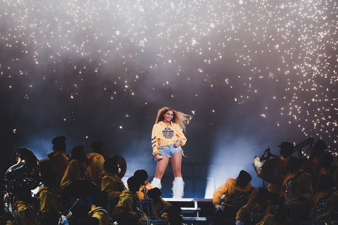 Beyoncé standing on stage in a yellow sweatshirt, denim cutoff shorts, and white fringe boots. She is holding a microphone in her right hand and has her left resting on her hip. There is a marching band sitting on risers below her all wearing matching yellow uniforms and holding their instruments. Behind Beyoncé there are sparks falling from the rafters above her.