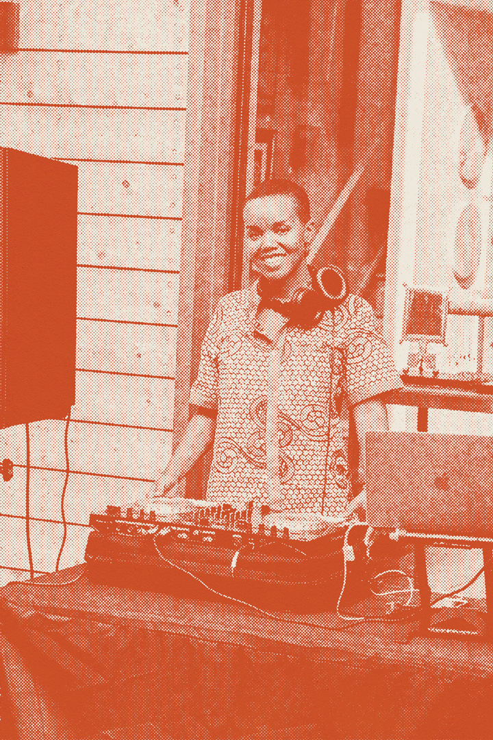 A person standing behind a DJ mixing booth wearing a patterned, button down shirt and a pair of large headphones hanging around their neck. The DJ mixing box is on top of a covered table and their is a macbook laptop on a stand next to the station. The photo has an orange halftone filter on top of it.