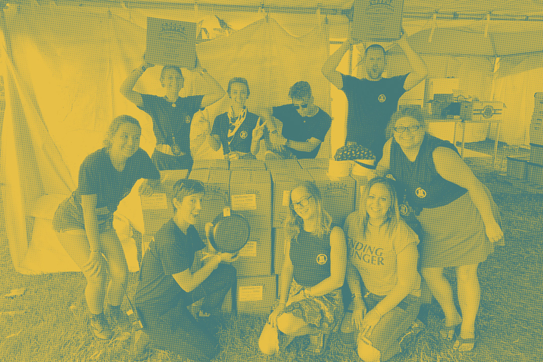 Small group of people smiling for photo in an outdoor tent. They are standing on grass and are surrounded by makeshift walls mades from handing tarps. They are circled up around a stack of medium-sized boxes and some of the people in the photo are holding the boxes above their heads. Many of the group are wearing matching shirts with a circular emblem in the upper left hand corner. Some are standing behind the boxes and other are squatted in front of them.
