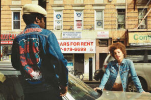 A city street with a man an woman posing for a photo in front of a car. The photo is from the 80s and has a vintage look to it. The man is facing away from the camera on the left side and the back of his jean jacket has handwritten graffiti letters on it. The woman is facing towards the camera on the other side of the car and is leaning on the hood. She has a small afro and is wearing a light blue jeans jacket over a white shirt. In the background is a moving car and a few storefronts that are selling cars, musical instruments, and other items.