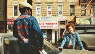 A city street with a man an woman posing for a photo in front of a car. The photo is from the 80s and has a vintage look to it. The man is facing away from the camera on the left side and the back of his jean jacket has handwritten graffiti letters on it. The woman is facing towards the camera on the other side of the car and is leaning on the hood. She has a small afro and is wearing a light blue jeans jacket over a white shirt. In the background is a moving car and a few storefronts that are selling cars, musical instruments, and other items.