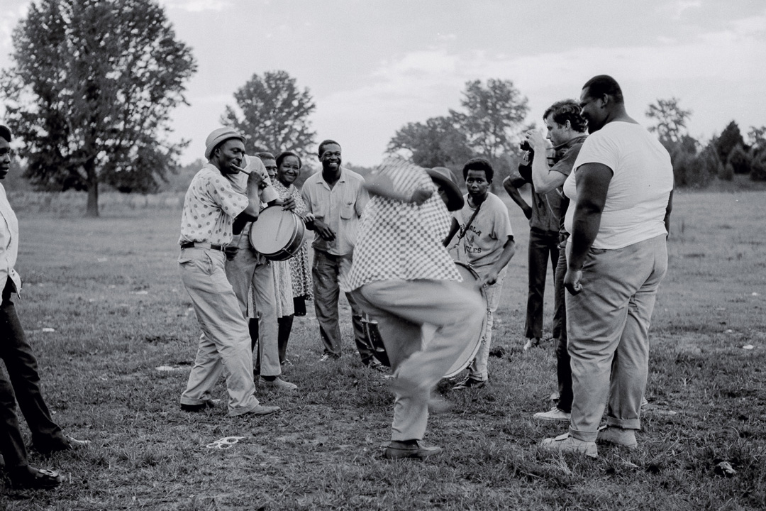 Black and white photo of a group of men and women dancing and playing music in a large open field. There are two marching band drums being played and one man playing a small fluted instrument. There is a man in the center in the middle of dancing so the image captured his motion blur. There is another man on the right side of the small group with a camera photographing the scene.