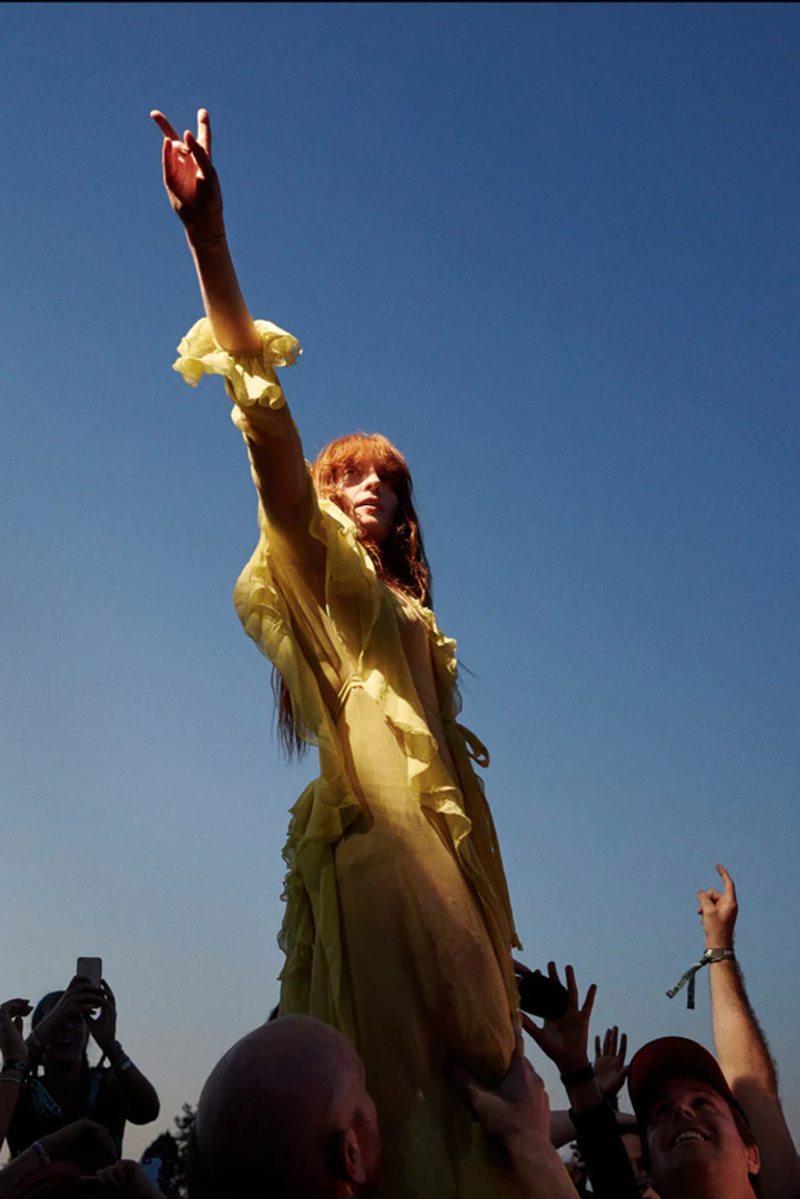 Woman in a long, flowy, yellow dress being held up by the legs over the crowd at a performance. She is holding up peace fingers towards the sky while hands reach up around her and someone sits on another person's shoulders holding up a phone.
