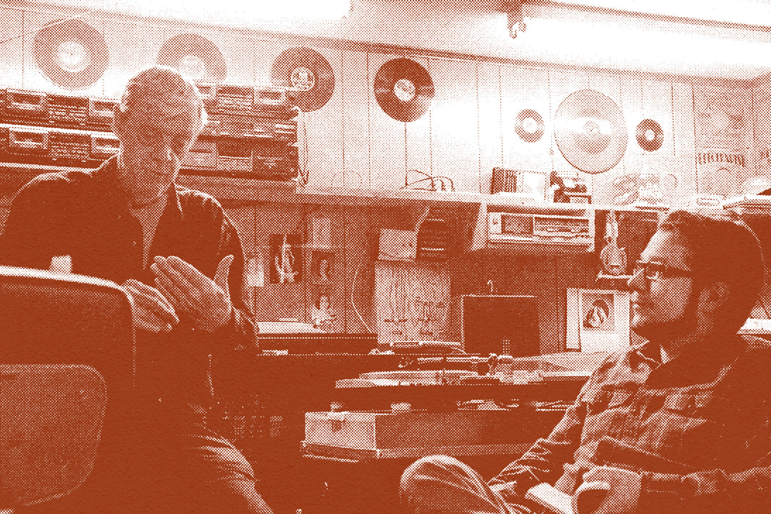 Inside of a small, wood paneled room where two men are sitting discussing. The walls are lined with shelves and topped with recording equipment, records, and other objects. The walls hve photos pasted on them and there is a large turntable in the center almost between the two men. The man on the left is older and is using his hands to describe something while the man on the right side watches. The image has a dark red-colored overlay and a halftone texture on them.