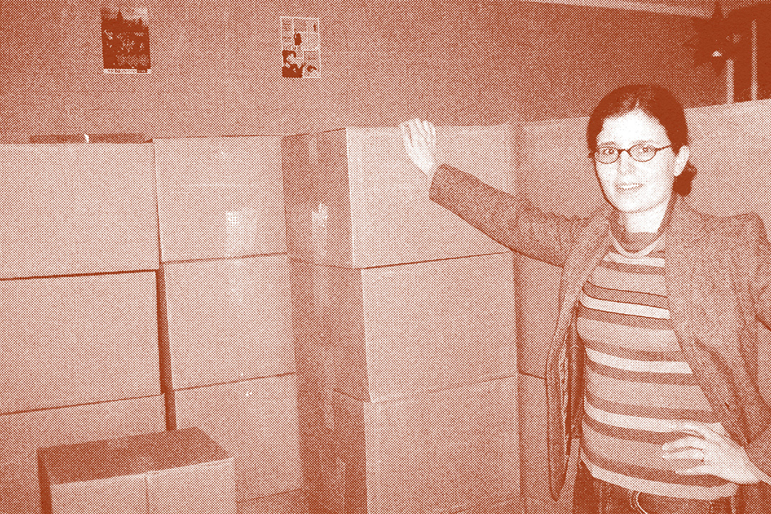 Woman posing for a picture in a small room filled with dozens of boxes. She is leaning with her right hand propped against the stack of boxes and her left hand on her hip. She is wearing pants, a striped shirt, blazer, and round glasses. The photo has a dark red-colored overlay on it and a halftone texture applied to it.