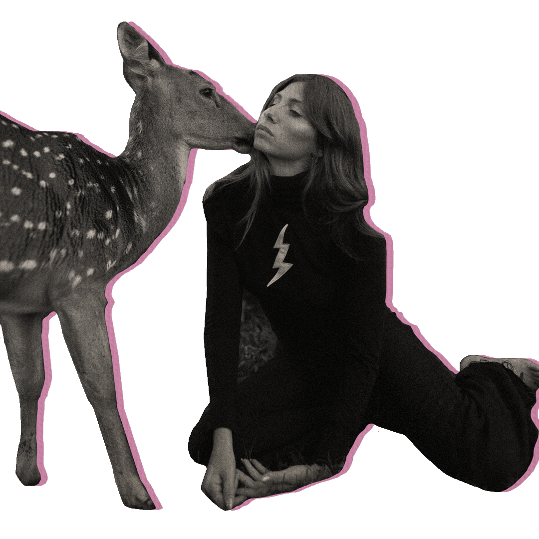 Black and white photo of a woman wearing black pants and a black long sleeve shirt with a lightning bolt on the front. She is sitting on the ground with her legs bent behind her while a deer stands next to her with its face touching her cheek. The two are cutout from the background of an image and have a pink-colored outline around them.