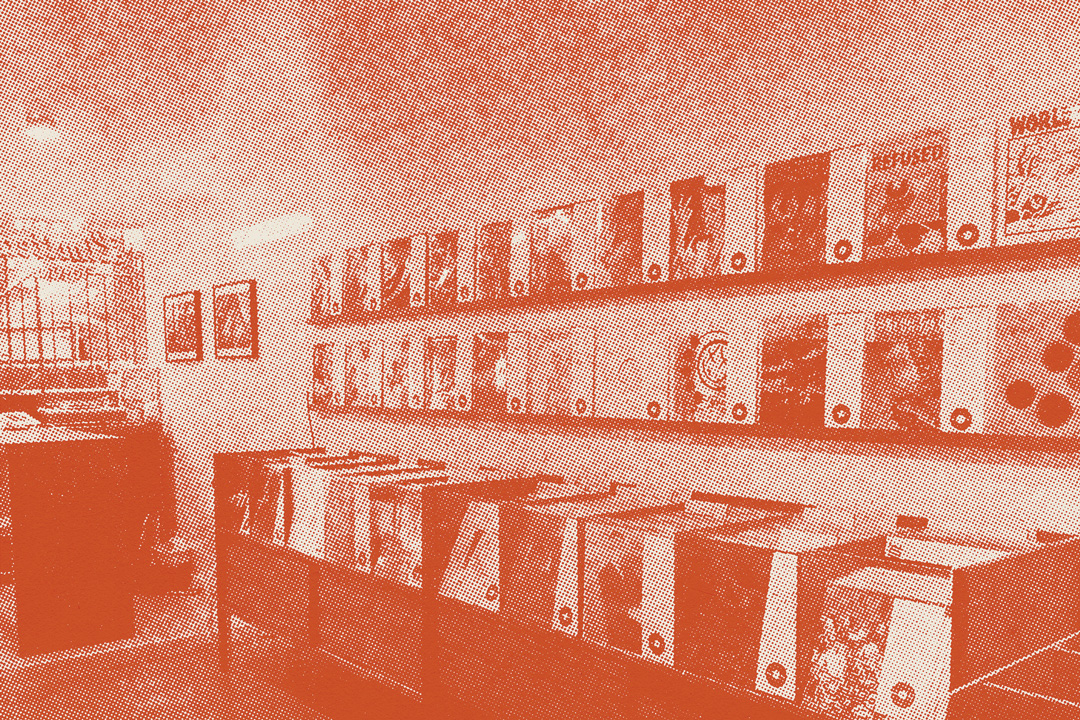 Inside of a small room that has two long shelves stacked on top of each other and are covered in records. Underneath the shelves is a long cabinet that is displaying stacks and stacks of records. To the side of the cabinet there is a desk and frames hanging on the wall. The photo has an orange halftone texture applied over it.