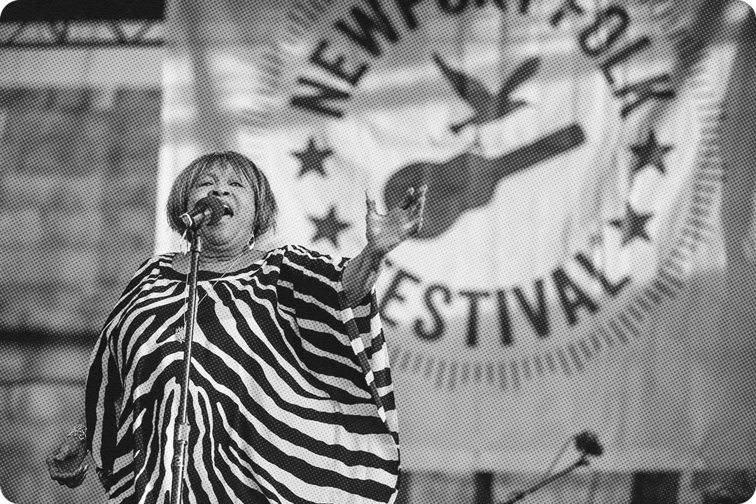 Black and white photo of Mavis Staples singing on stage. Staples is singing in front of a microphone on a stand while raising her left hand upwards towards the audience. She is wearing a large flowing dress in a zebra print pattern. Behind her on stage is a big banner for the Newport Folk festival with the organization's logo on it.