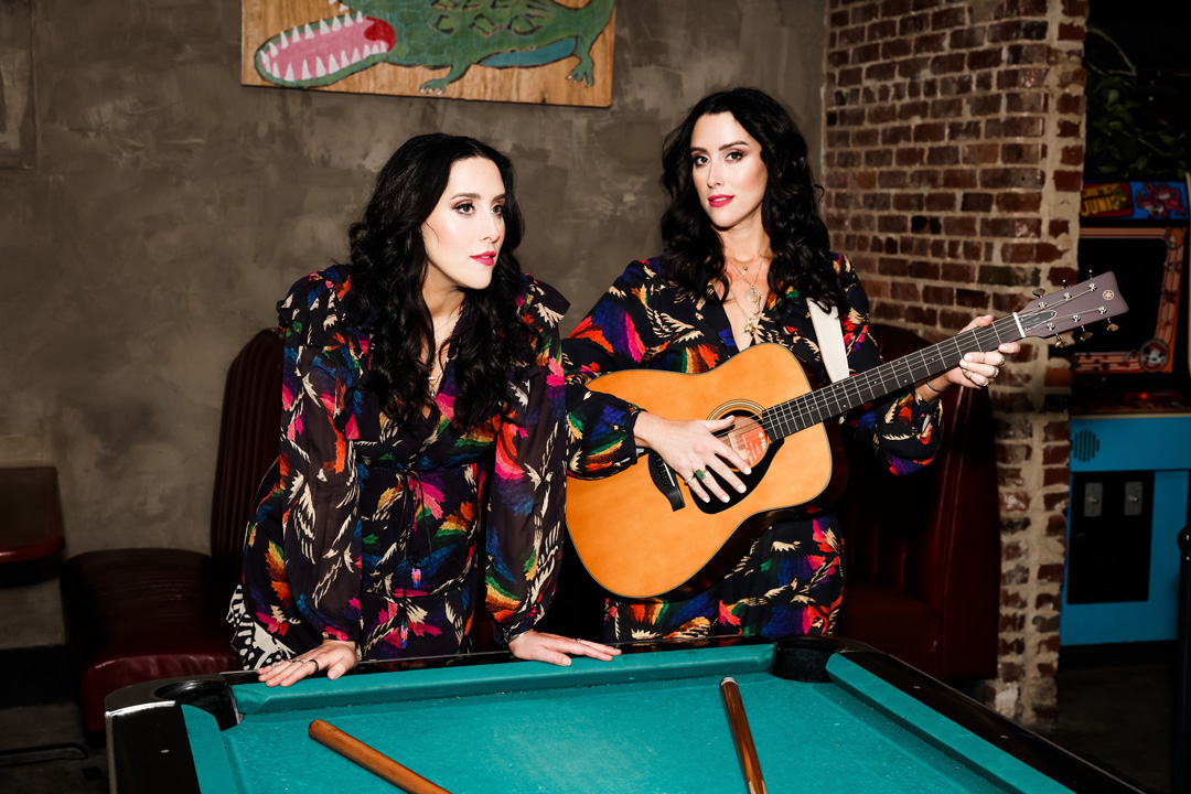 Two women pose for a photo leaning up against a green pool table. They are standing in a room that has brick and concrete walls, arcade games, and leather chairs in the background. The women both have long, dark, wavy hair and are wearing matching black and rainbow colored outfits. The woman on the right is holding an acoustic guitar and the other is leaning on the table.