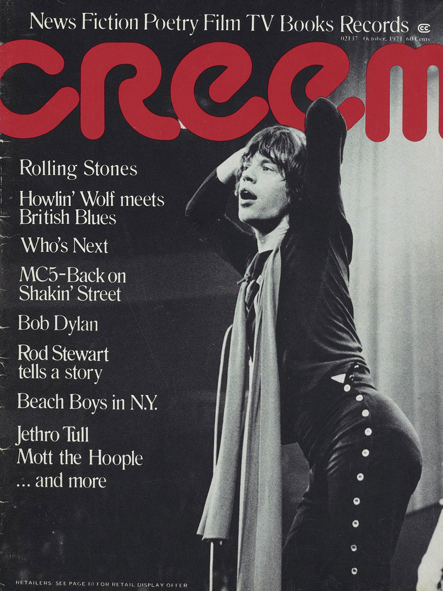 Photo of CREEM's October 1971 cover featuring an action shot of Mick Jagger. The cover is black and white and Mick Jagger is wearing a black shirt, a long scarf tied around his neck and black pants with buttons up the length of each pant leg. He has his hands behind his head and is singing out to an unseen crowd.