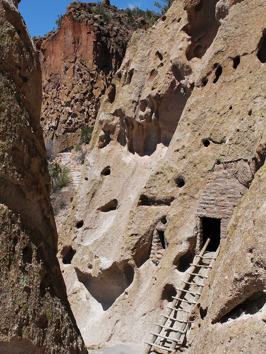 Photo of one of the Pueblo dwellings in one of the canyons at The Bandelier National Monument in Santa Fe, New Mexico. Amidst a large mass of light brown sandstone rock, there is a small rectangular black opening in the side of the mountain with what appears to be a hand-built wooden ladder reaching from the opening to the floor of the canyon.