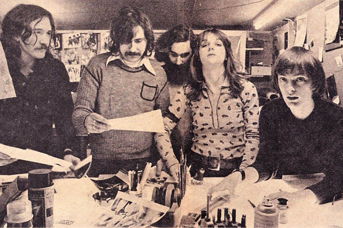 Grainy, black and white photo of original CREEM staff members standing in a row in front of a work desk. There are 3 men to the left looking at a photo held by the middle man, a woman in the middle looking at the camera and a man with a bowl cut sitting at the very end of the row of people, looking up at the camera.
