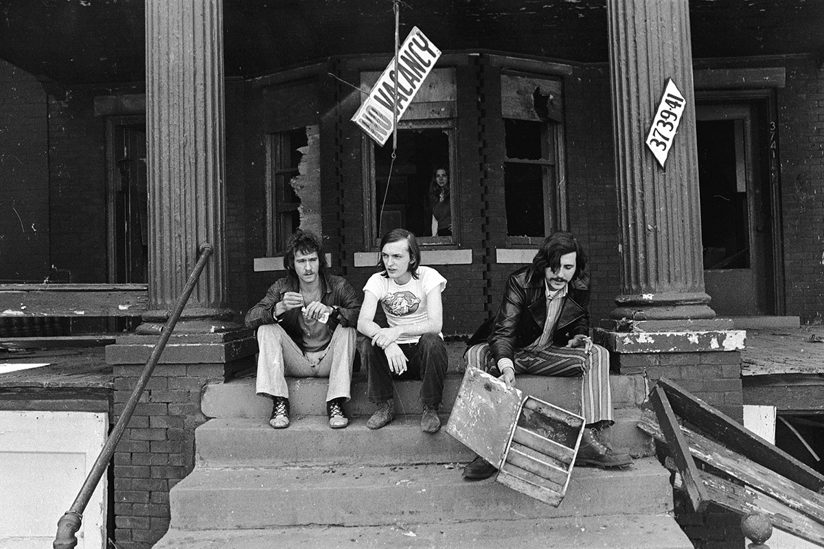 Black and white photo of Barry Kramer, Dave Marsh and Lester Bangs outside on the concrete steps of CREEM's old Detroit offices. All three men have long hair and are dressed in '70s-style clothing. Lester Bangs appears to be sorting through some left over furniture parts on the right. A woman can be seen looking out of the window of the building behind the stairs. There is a "NO VACANCY" sign hanging haphazardly from a cable above the stairs that the men are sitting on. 