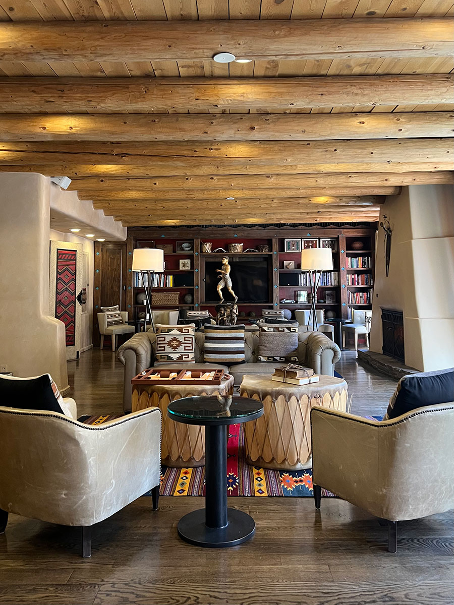Photo of the lobby of The Inn of the Anasazi. The entire room is decorated in southwestern style decorations and has a light brown tone to it, from the two leather arm chairs seen at the front of the photo to the wooden ceilings. A large floor-to-ceiling bookshelf lines the back wall and in front of it there is a golden statue of a person running. There is a fireplace on the right wall which is an off-white color. 