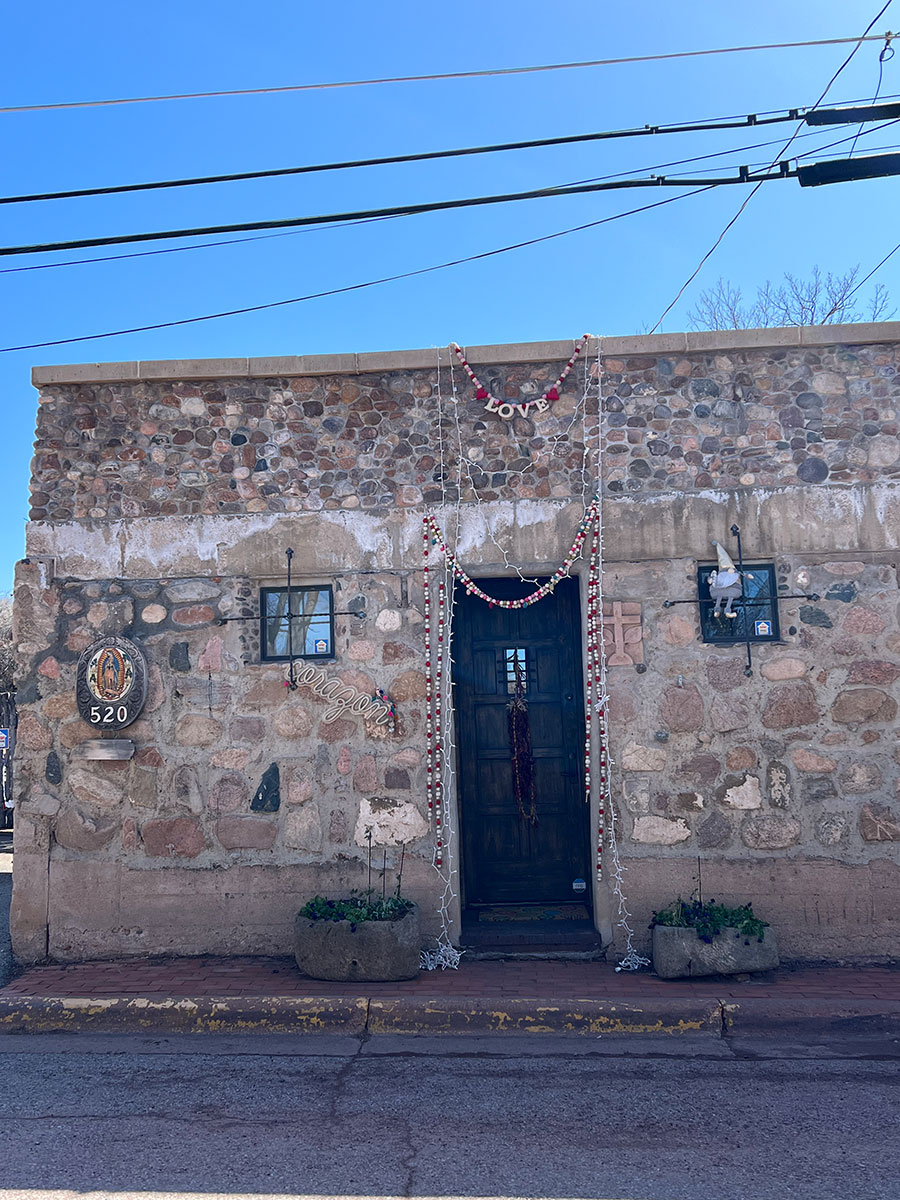Photo of a small building made of various sized stones on Canyon road. A dark brown door with a rostra (a bushel of dark red dried Chile peppers) is hanging from the center of the door and red and white garland is hanging above and on either side of the doorframe. The word "LOVE" is in the center on the top strand of garland.