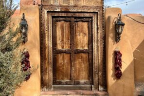 Photo of a large old brown wooden door embedded into the side of an orange-brown cement wall in Santa Fe, New Mexico. There is an old lamp either side of the door. Dark red bushels of ristras (bushels of dried chile peppers used for decoration in New Mexico) are hanging from each lamp.