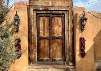 Photo of a large old brown wooden door embedded into the side of an orange-brown cement wall in Santa Fe, New Mexico. There is an old lamp either side of the door. Dark red bushels of ristras (bushels of dried chile peppers used for decoration in New Mexico) are hanging from each lamp.