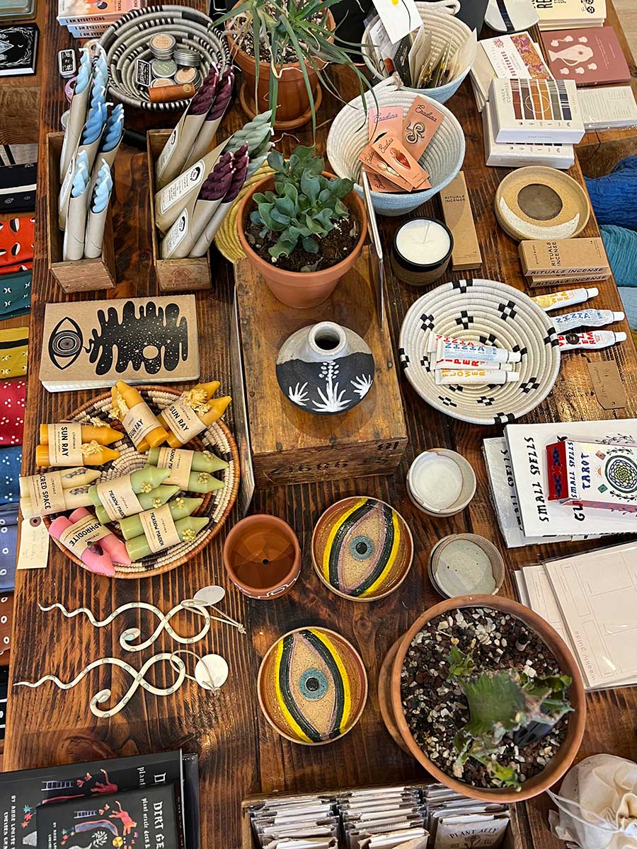 Aerial photo of a wide array of items and trinketsbeing sold on a wood table at a store in Santa Fe, New Mexico. There are handmade coasters with large outlines of eyes on them, small pots decorated in Southwestern patterns, potted plants, a bowl of pink, green and yellow candle sticks, tubes of paint, small pocket-sized journals and packs of tarot cards.