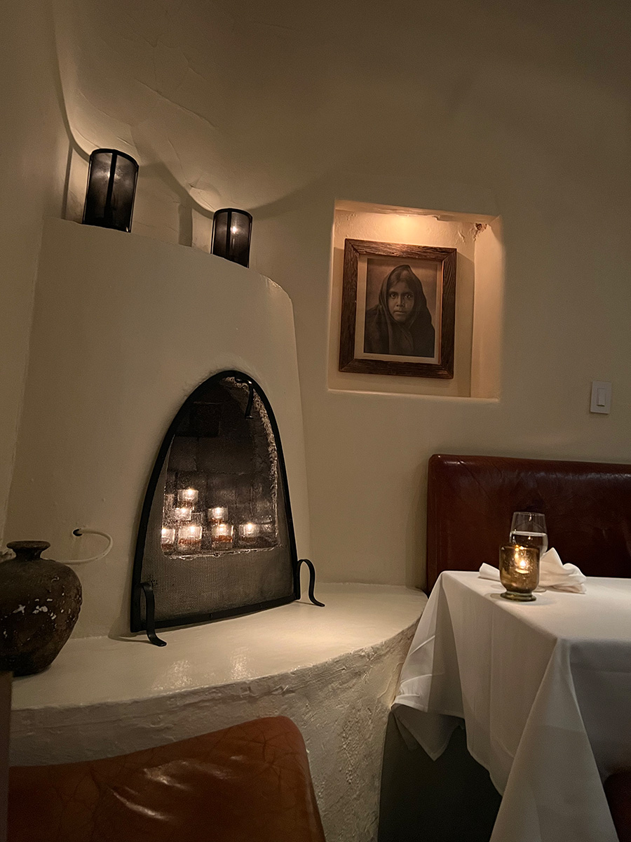 Photo of a small fireplace and booth table at the Geronimo restaurant in Santa Fe, New Mexico. The room is very dim and lit only by the candles stacked in the small curved fireplace and the two candles sitting on the ledge above it. The walls are an off-white sandstone. There is a black and white portrait set into the wall of a young woman wearing a scarf around her head and staring at the onlooker. Part of the booth dark red booth chair and white table cloth can be seen on the left side of the photo. 