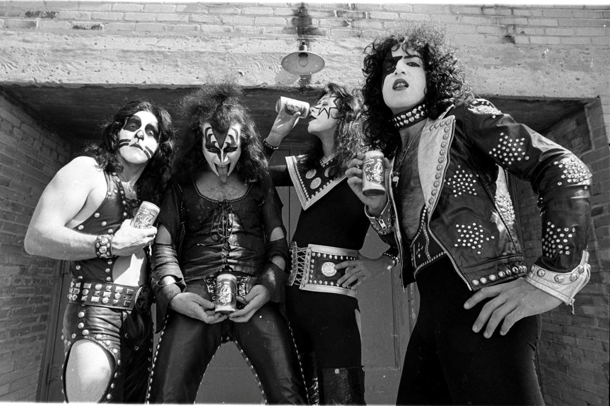 Photo of the four members of the rock band Kiss taken for CREEM magazine. Each member is decked out in black and white face paint, long, curly black hair and leather jackets and jumpsuits with decorative metal studs. They are each holding a can with the CREEM magazine logo on it.
