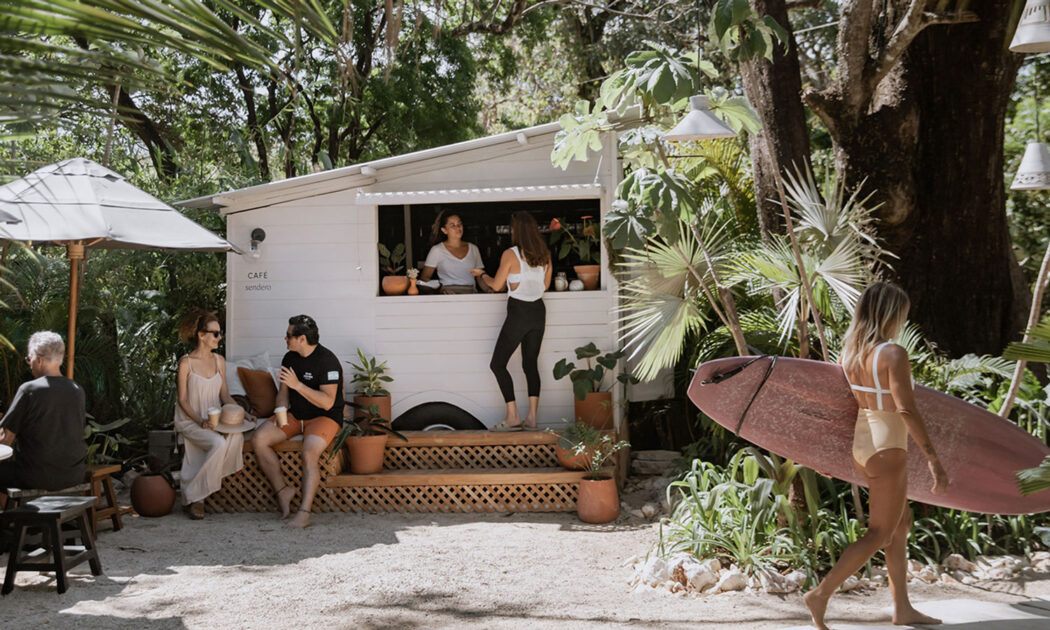 Photo of a person getting coffee at CAFE Sendero. The cafe is a small white wooden bungalow with a service window. People are sitting on couches under umbrella in conversation to the left. A woman in a pale yellow one piece swim suit is walking away with a light pink surf board under her arm in the righthand side of the photo.