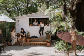 Photo of a person getting coffee at CAFE Sendero. The cafe is a small white wooden bungalow with a service window. People are sitting on couches under umbrella in conversation to the left. A woman in a pale yellow one piece swim suit is walking away with a light pink surf board under her arm in the righthand side of the photo.
