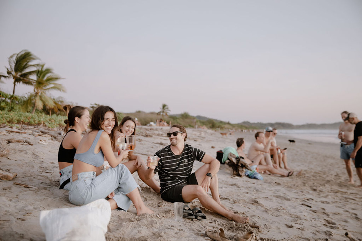 Photo of a group of people lounging together on the sandy beach in Nosara, Costa Rica at sunset. People in the group have glasses of rose in their hands and are toasting. One person sitting on the sand is turning back and smiling at the camera.