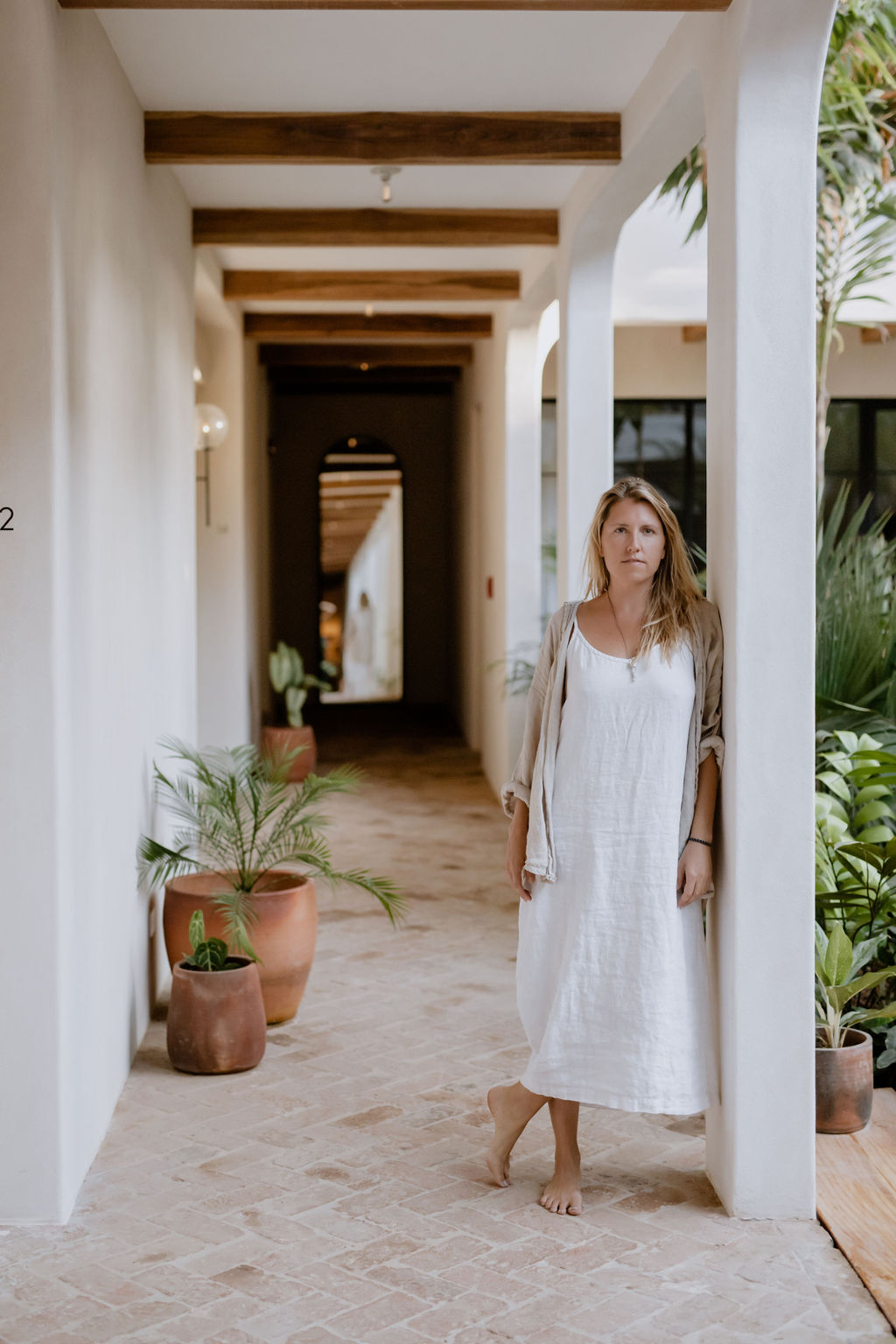 Photo of Stephanie Tannenbaum leaning against a white pillar in a serene outdoor hallway wearing a flow white dress and no shoes.
