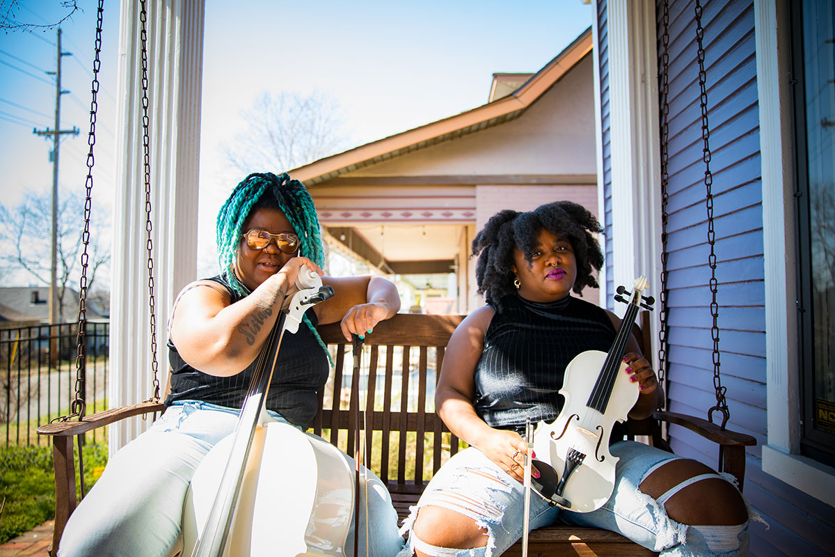 Photo of SistaStrings duo Chauntee Ross (left) and Monique Ross (right). The two sisters are sitting next to each other on a swinging porch bench wearing black sleeveless mock neck tops and light wash jeans. Each sister is holding their personal instrument; Chauntee is holding the top of her white cello and Monique is holding her violin in her hands on her lap.
