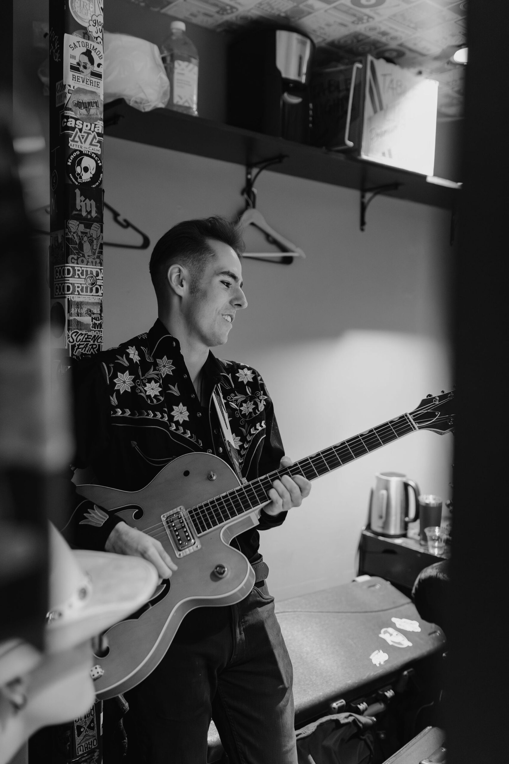 A black and white picture of a man tuning a guitar in a green room backstage