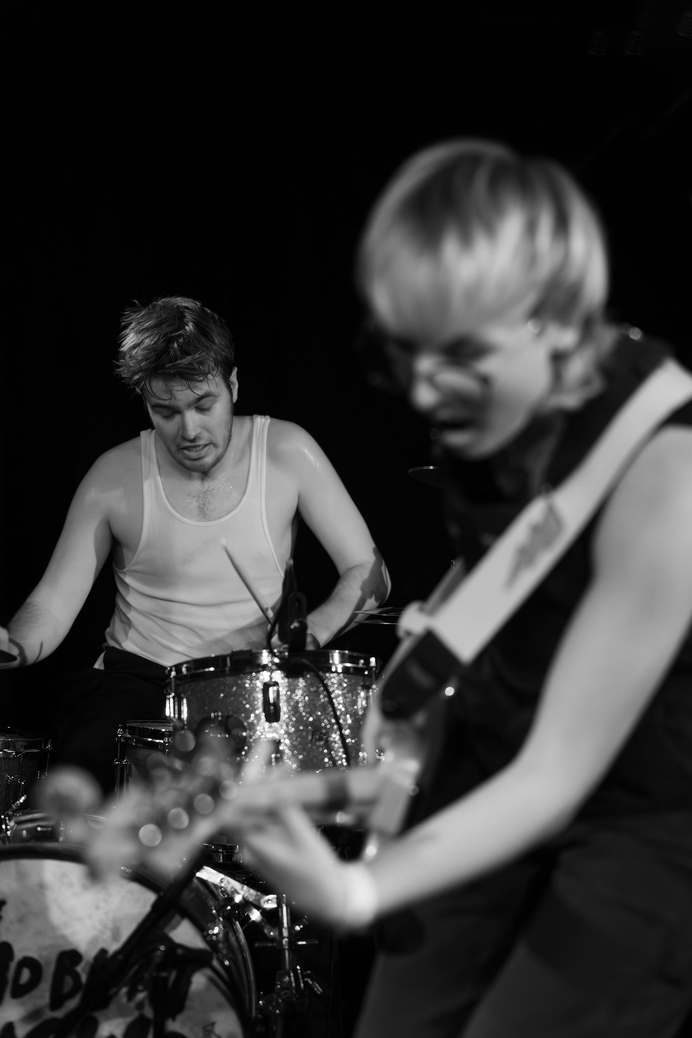 A black and white photo of a woman playing bass in the foreground with a drummer in a tank top in the background