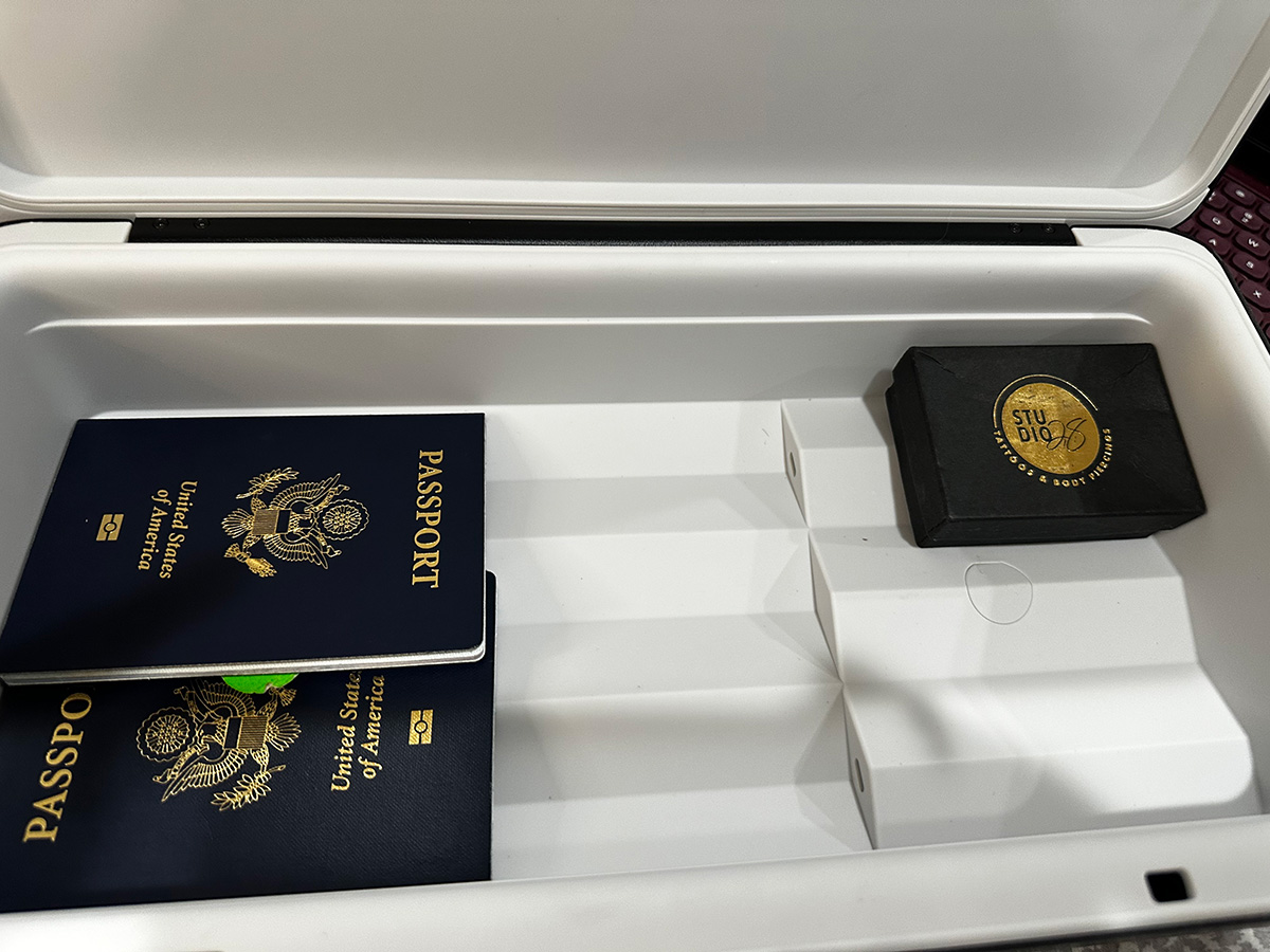 Close up photo of the inside of Monique Ross's TROVA safe. The inside of the safe is light gray. In the bottom compartment to the left there are two rectangular, dark navy blue United States of America passports. In the right corner, they're a small black box with a golden emblem holding Monique's nose ring.