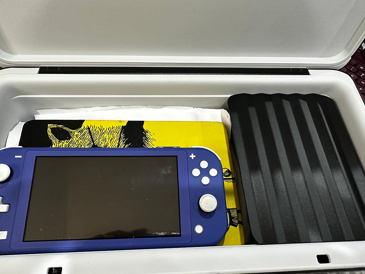 Close up photo of the inside of Monique Ross's TROVA safe. The inside of the safe is light gray. The items stored inside the safe include a bright yellow notebook filled with loose papers, a royal blue Nintendo Switch and a  black cover concealing the other items in the safe on the right.
