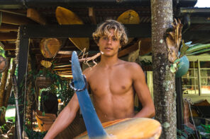 Close up photo of Rasmus King in front of his rustic cottage, shirtless and carry9ing a blue and yellow surf board.