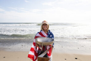 Photo of Caroline marks standing on the beach with a baseball cap on and wrapped in a red, white and blue American flag. Caroline is holding the large silver and gold cup/trophy that she won at the WSL World Championships. She is smiling big at the camera.