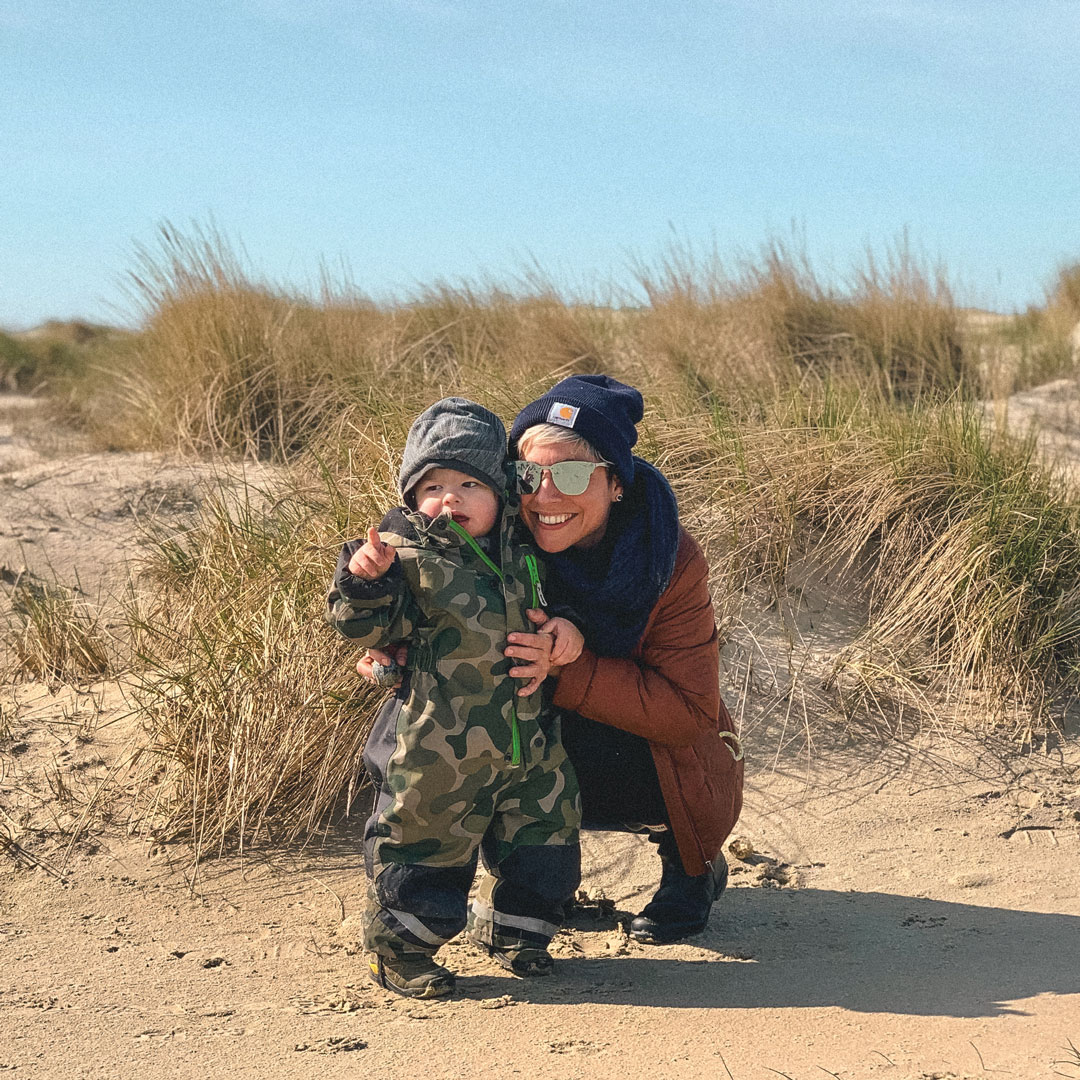 A women bends down beside a toddler. Both are wearing beanies and cold weather clothing. They stand on a beach in front of low dunes and tall grass. 