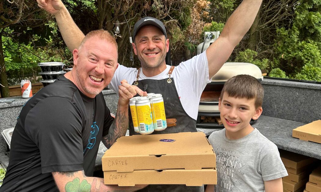 Photo of Joe Prota with his arms raised (middle) and a man and his son from Dad's Night on either side of Joe, smiling for the camera. The dad is holding two light brown cardboard pizza boxes and a four-pack of yellow beer cans. The little kid is on the other side of Joe, smiling.