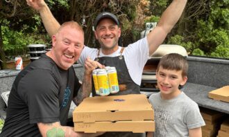 Photo of Joe Prota with his arms raised (middle) and a man and his son from Dad's Night on either side of Joe, smiling for the camera. The dad is holding two light brown cardboard pizza boxes and a four-pack of yellow beer cans. The little kid is on the other side of Joe, smiling.