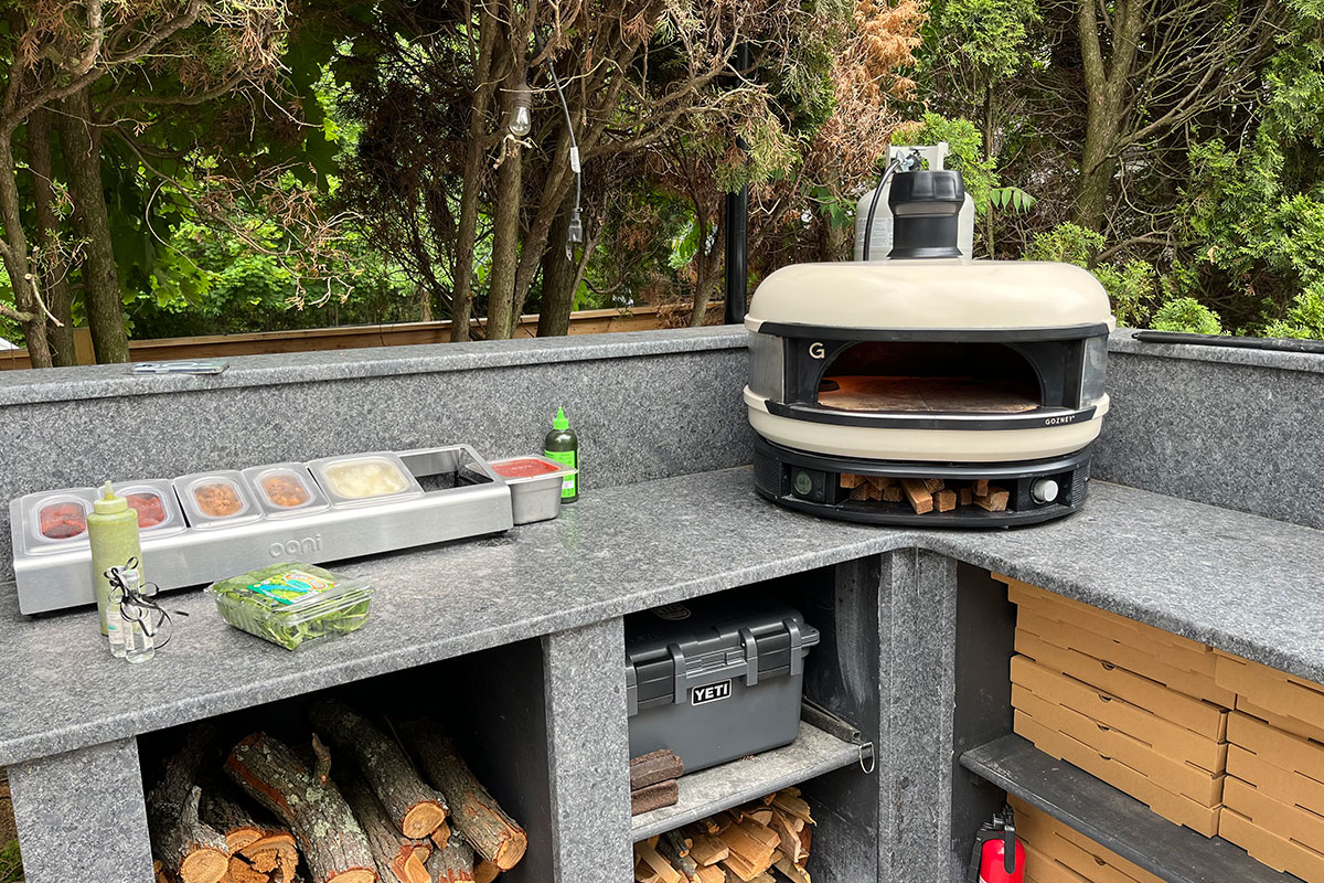 Photo of Joe's setup for making pizza. There is an outdoor gray marbled bar with condiments on the left and a medium-sized wood-burning pizza oven in the corner. In the storage cubbies under the bar, there are piles of would, dozens of light brown cardboard take-out pizza boxes, a bright red fire extinguisher and a small dark gray yeti cooler.