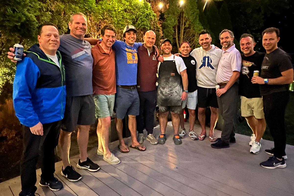 Group photo of a bunch of dads at Joe's Pizza Night event posing with their arms around each other. It is dark outside except for the lanyard of outdoor lights lining the patio. Joe is in the center of the group wearing his once-black apron covered in white flour.